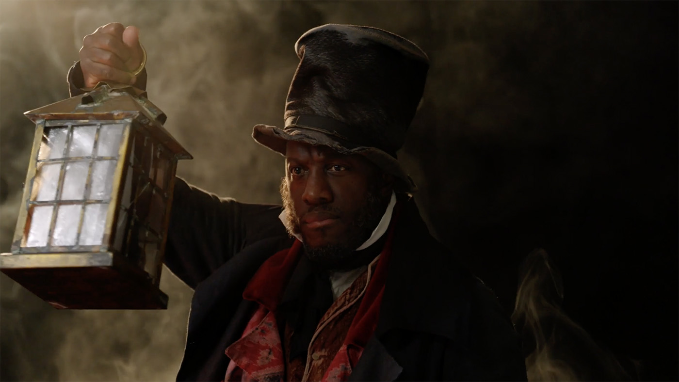 Actor Giles Terera emerges from a misty dark background. He's holding a lantern with a warm orange glow. He's wearing a crumpled top hat, a dark overcoat and a shabby waistcoat.