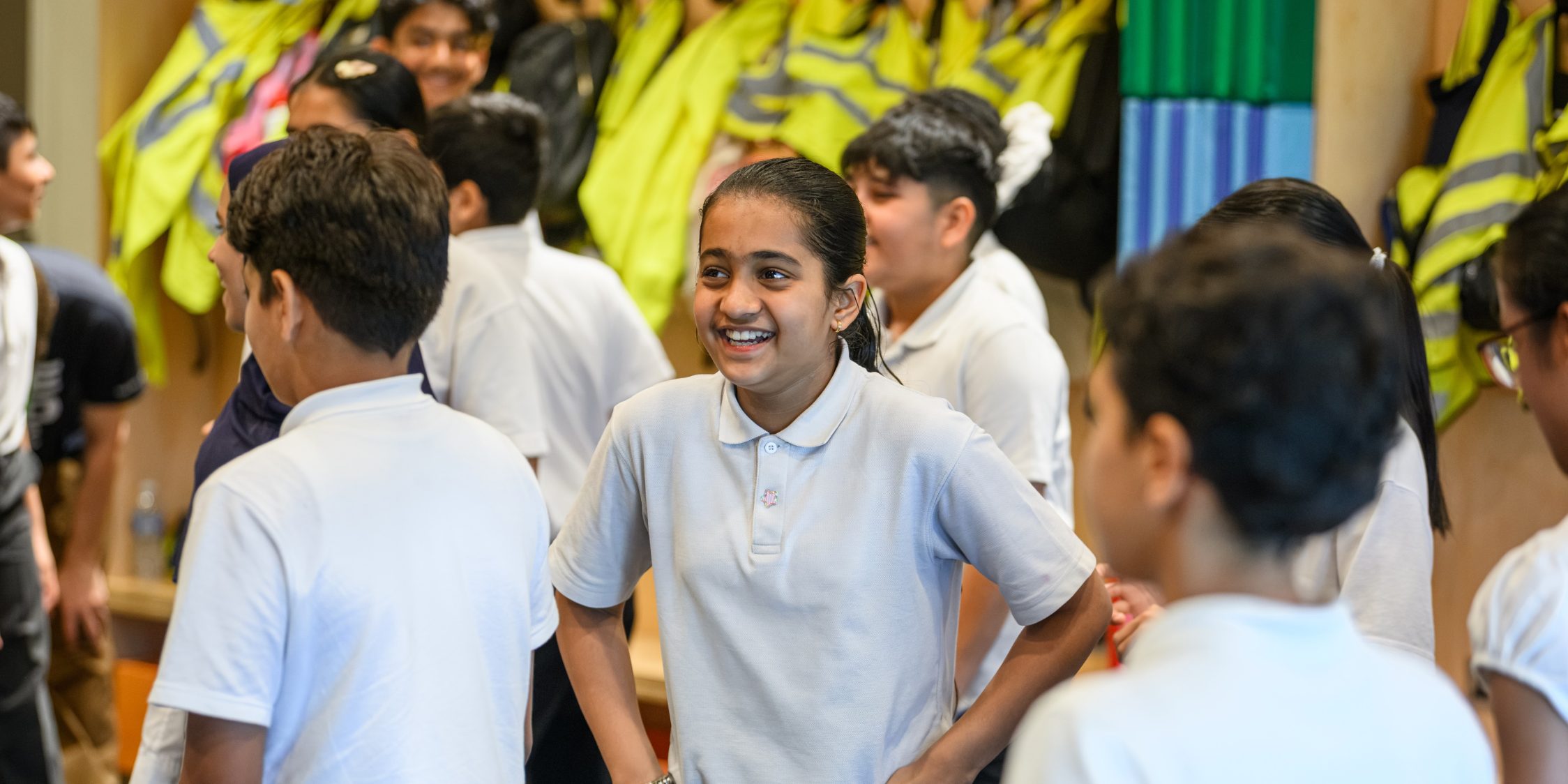 A group of school children are standing together looking in different directions. In the centre, a girl is laughing with her hands on her hips.
