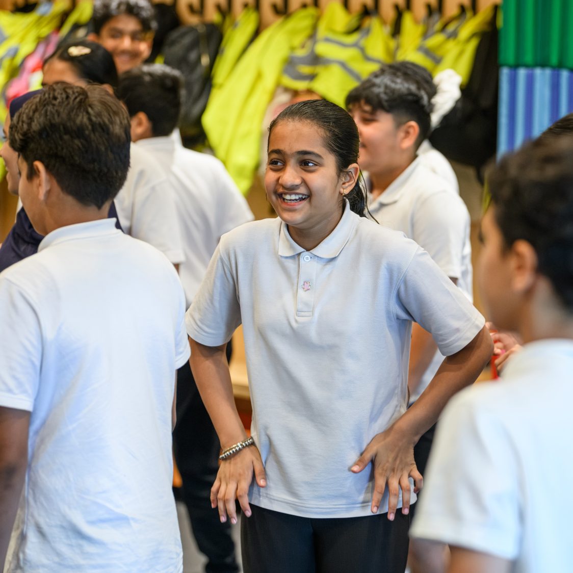 A group of school children are standing together looking in different directions. In the centre, a girl is laughing with her hands on her hips.
