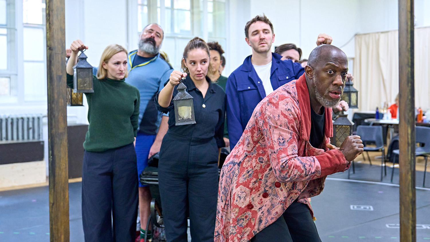 Giles Terera wears a tattered red dressing gown as he cautiously cowers. He's flanked by members of the cast of A Christmas Carol.