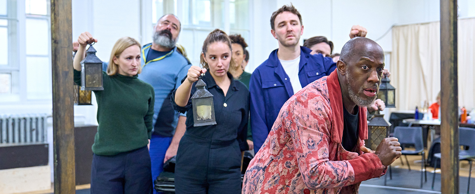 Giles Terera wears a tattered red dressing gown as he cautiously cowers. He's flanked by members of the cast of A Christmas Carol.