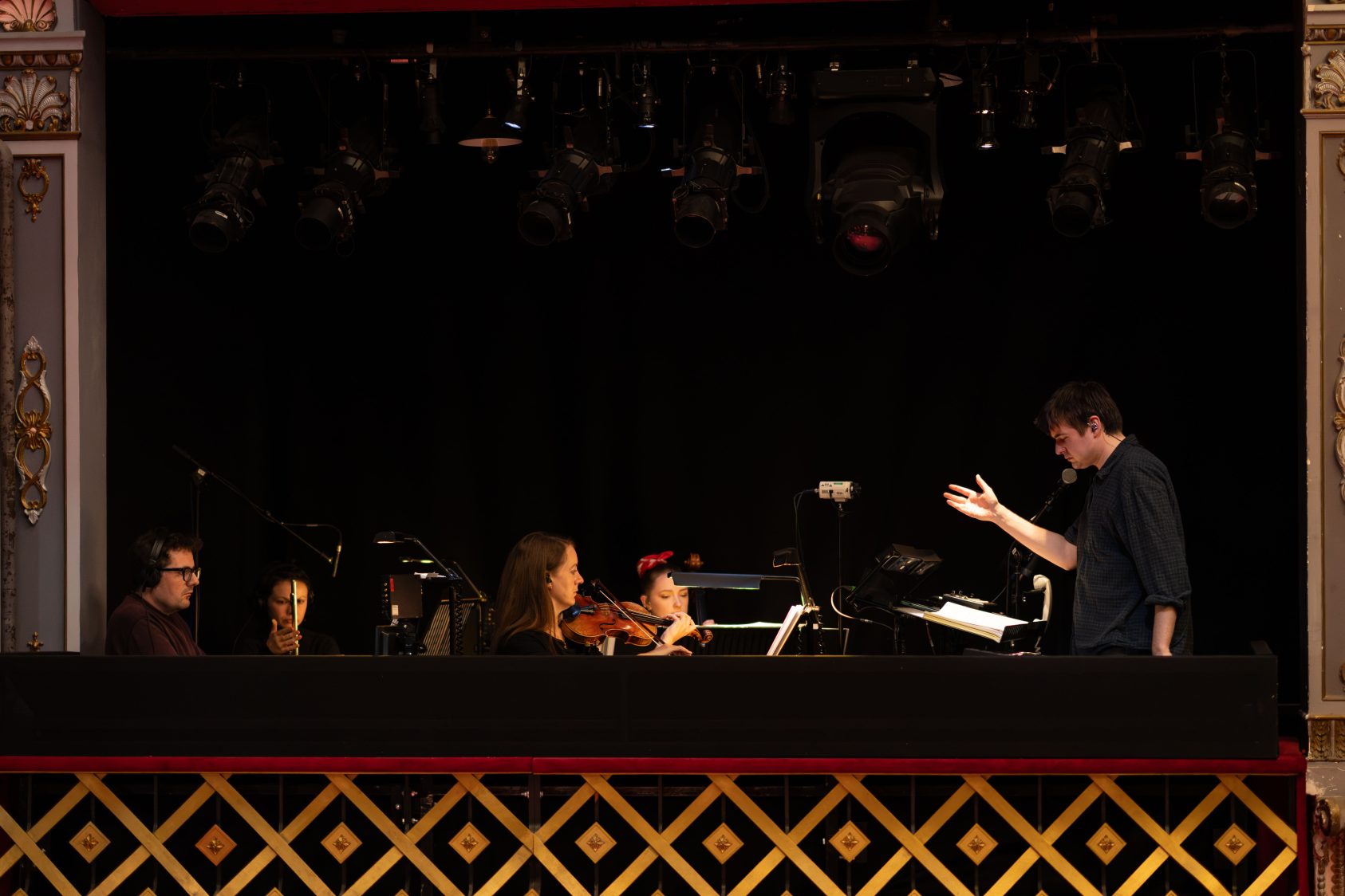 A Musical Supervisor from Martin Guerre stands in one of the ornate gilded boxes in The Old Vic auditorium conducting the assembled musicians.