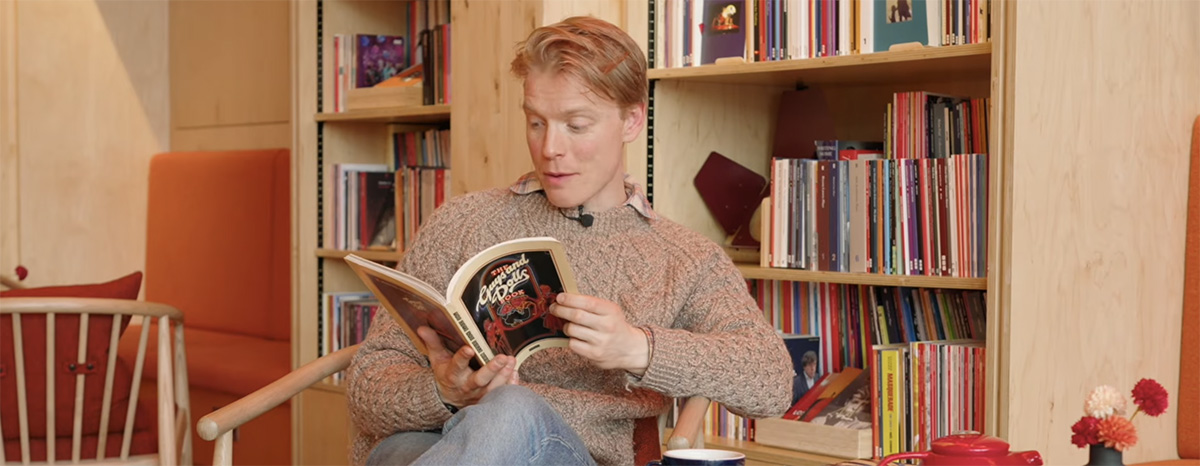 Freddie Fox sits on a chair in front of a shelf of books reading a copy of 'Guys and Dolls'