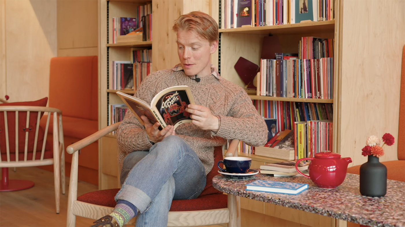 Freddie Fox sits on a chair in front of a shelf of books reading a copy of 'Guys and Dolls'