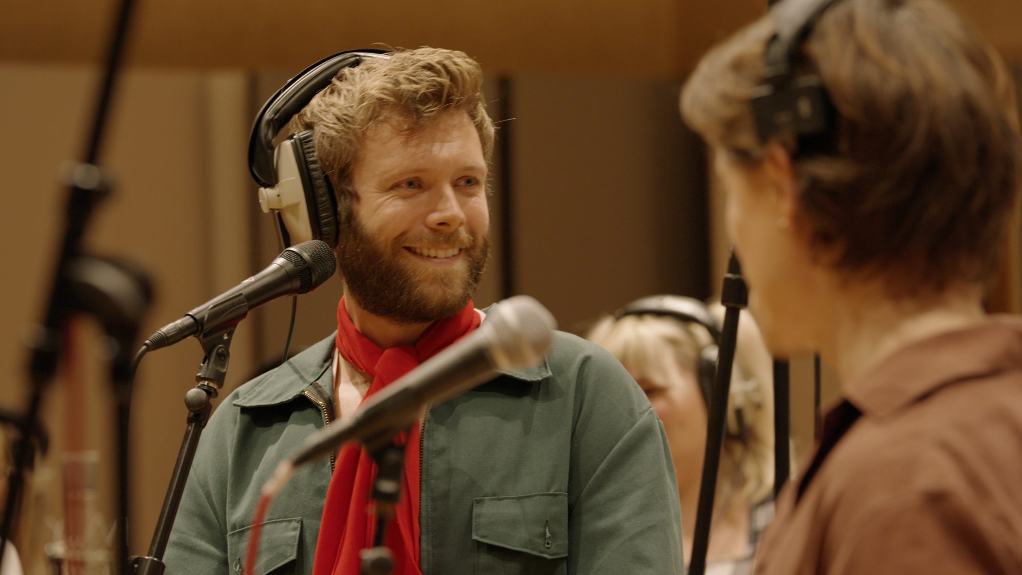 Actor Shane O'Riordan smiles as he stands in a recording studio, surrounded by microphones. He wears large headphones and faces another cast member, who has their back to us.