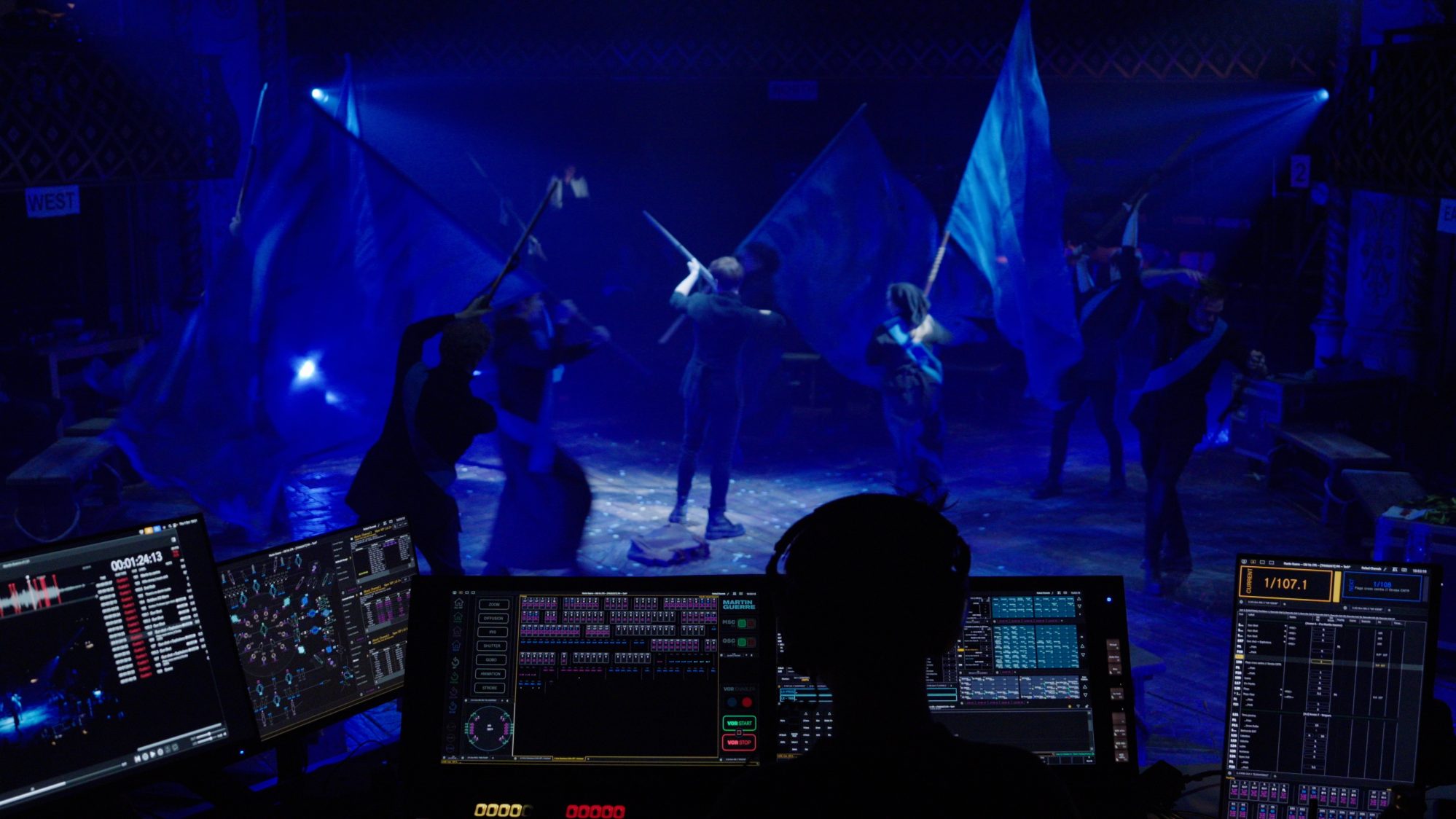 A view from behind the sound desk during a rehearsal, with illuminated computer screens in the foreground and the cast of Martin Guerre waving blue flags in the background