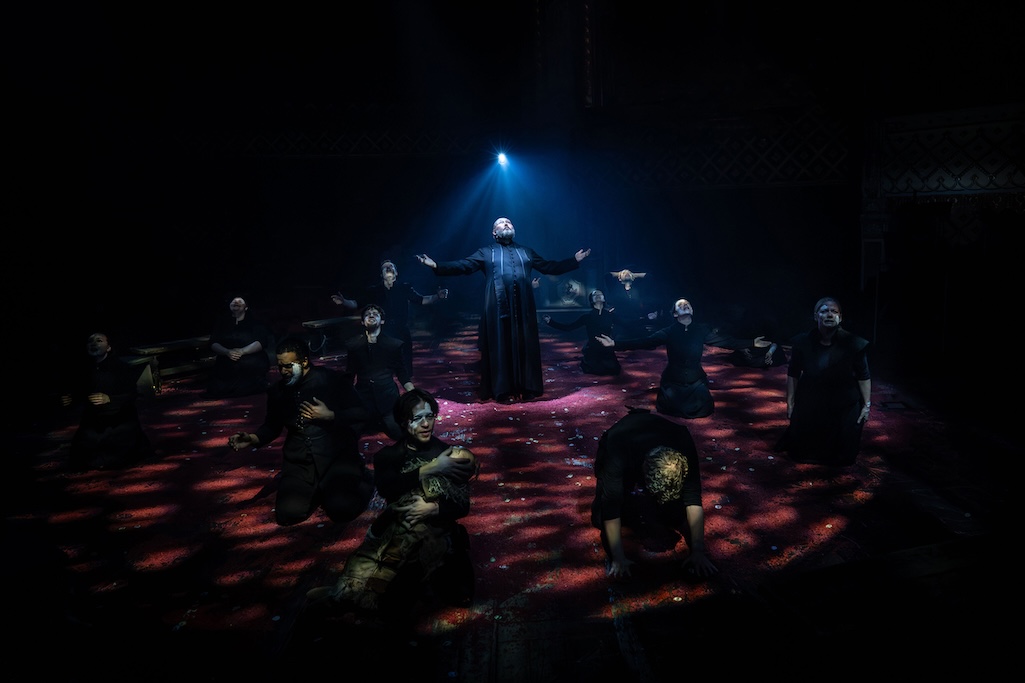 The cast of Martin Guerre kneel in a circle with arms outstretched and praying heavenward, bathed in dappled light.